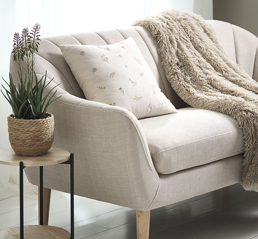 RASMUS konstgjord växt i STEFAN växtkruka, AGERTIDSEL-kudde och MYRFIOL-kast Konstgjord aloe vera-växt med blommor, broderad kudde och fuskpäls på beige soffa