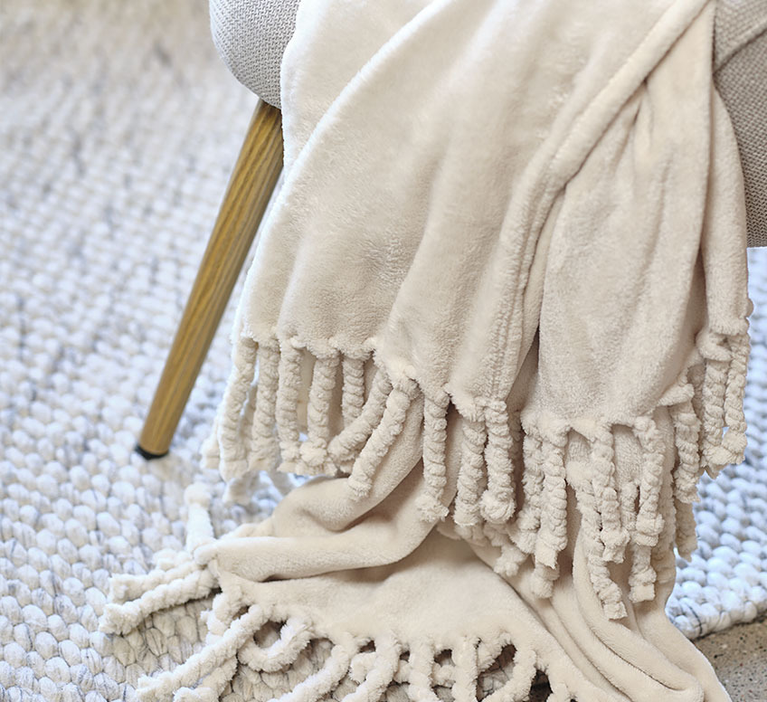 BONDEROSE pläd i beige kulör. En beige filt som ligger över en stol som står på en ljus vävd matta.