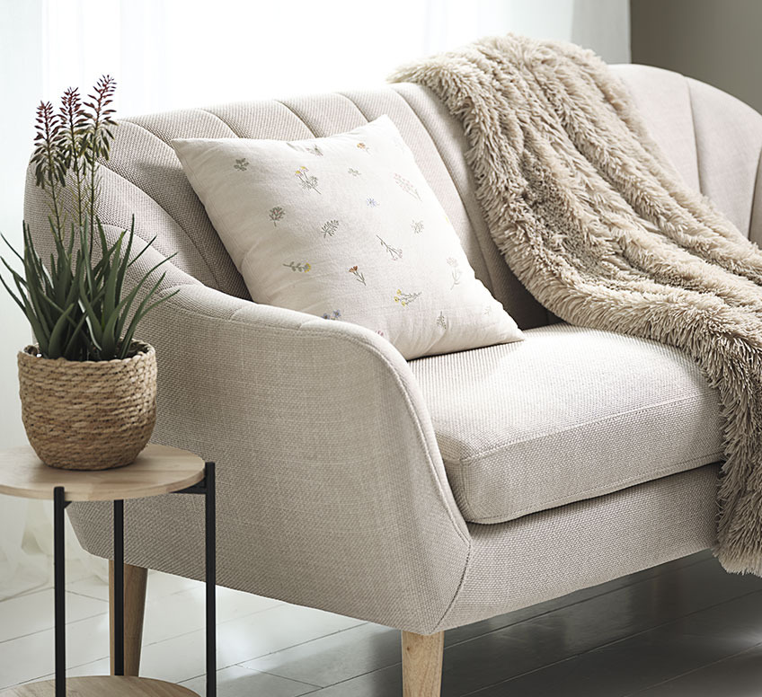 Benvit soffa med en filt ovan på med en kudde och ett sidbord med en växt. 