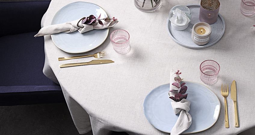 Uppdukat bord med ljusblå tallrikar, guldiga bestik och rosa glas på en beige linneduk 