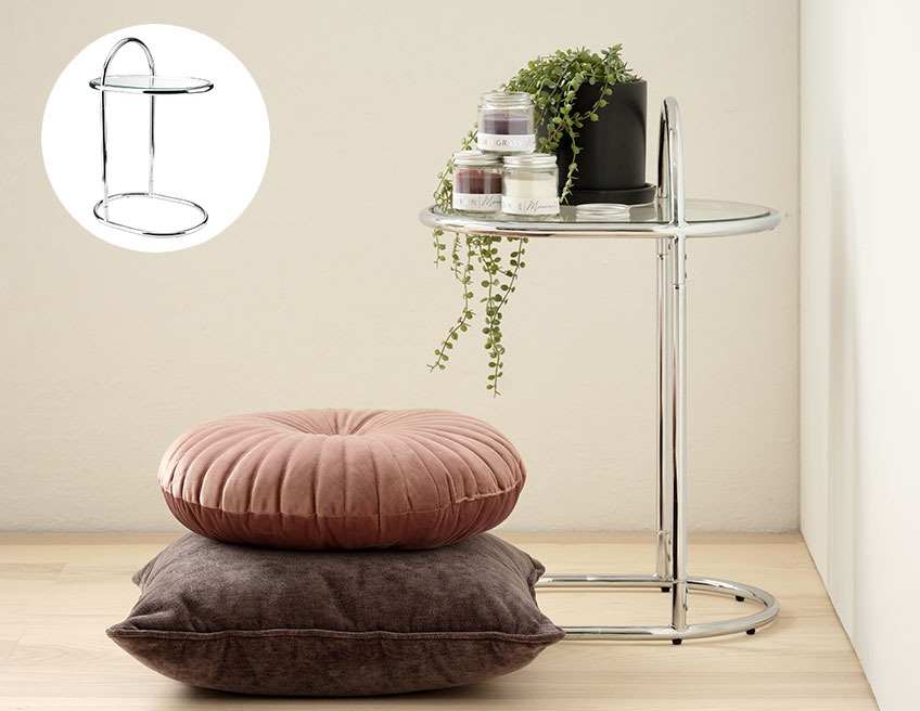 CURVEY sidobord i krom och glas med AXEL växtkruka på Ett kromat sidobord med en krukväxt i ett hörn och två kuddar på golvet