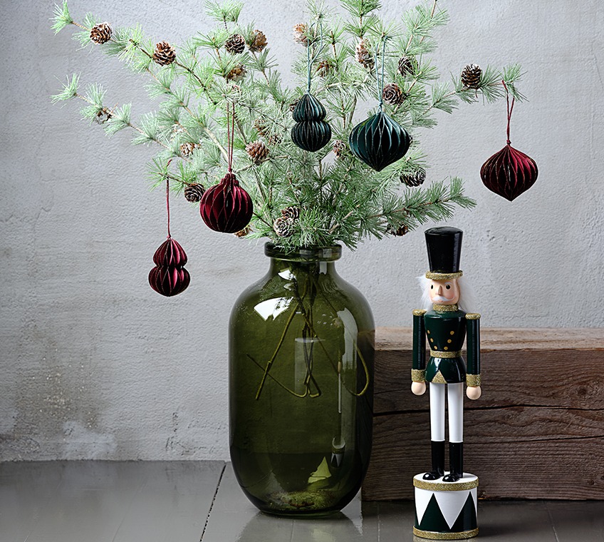 Grön glasvas fylld med konstgjorda grenarr med kottar, dekorerad med julpynt i grönt och rött. Bredvid star en julfigur. 