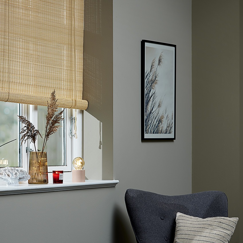 Rullgardiner av bambu vid ett fönster med en vas, ett doftljus och en batterilampa i fönsterbrädan