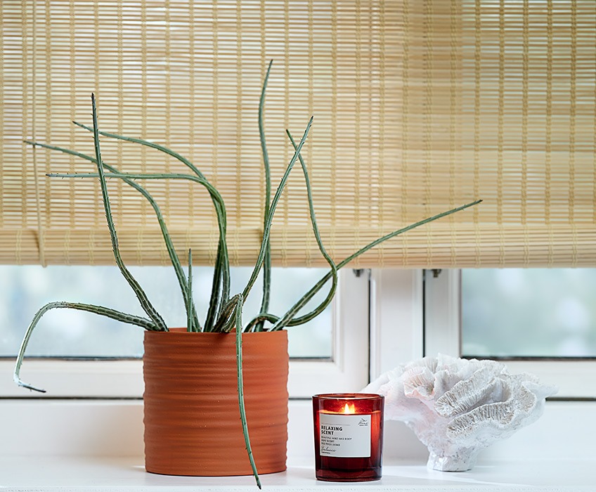 Rullgardiner av bambu i ett fönster med en krukväxt, ett doftljus och en prydnad i fönsterbrädan