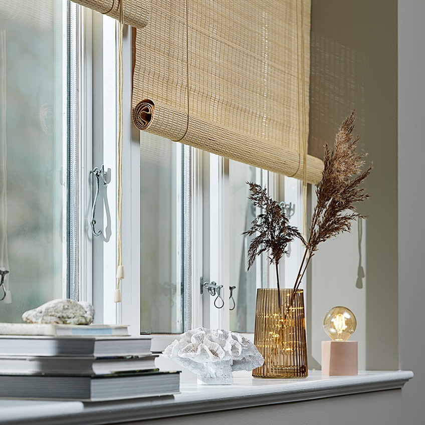 Rullgardiner i bambu i ett fönster med växtkruka, ornament, vas, doftljus och batterilampa i fönsterbrädan