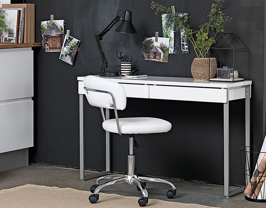 Litet vitt skrivbord med lådor och en vit kontorsstol vid en svart vägg