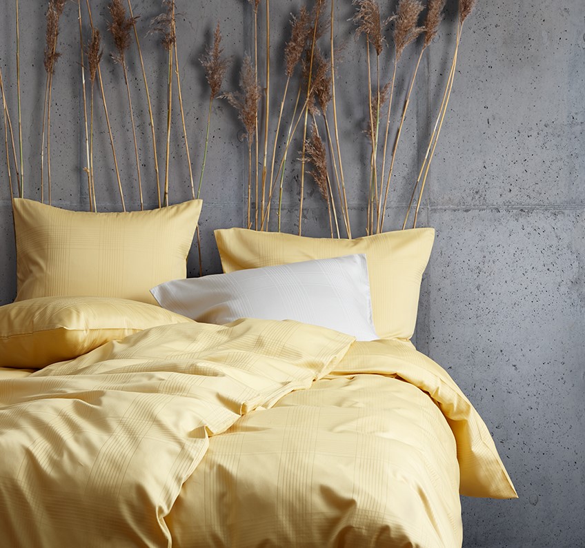 Gult sängkläder av 51% återvunnen polyester och 49% bomullssatin i ett sovrum med betongväggar