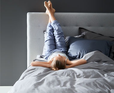 En kvinna ligger i en säng med benen upp mot sänggaveln bland kuddar och ett täcke. 